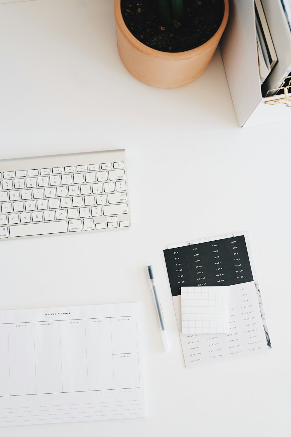 Overhead view of a wireless keyboard, planner, and plant on a white desk.